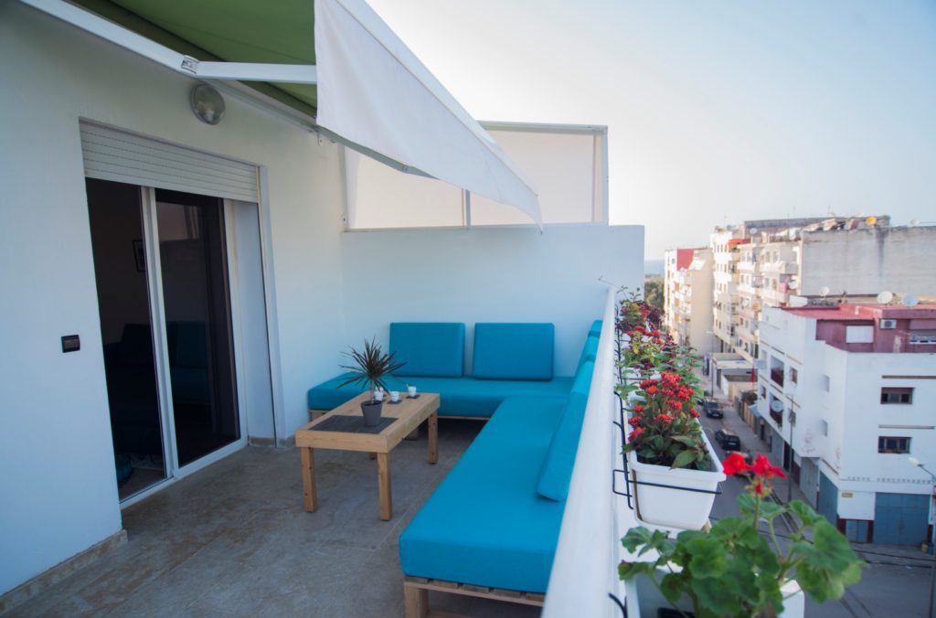Modern apartment balcony with teal blue armchairs, a small table and potted plants, city view in the background