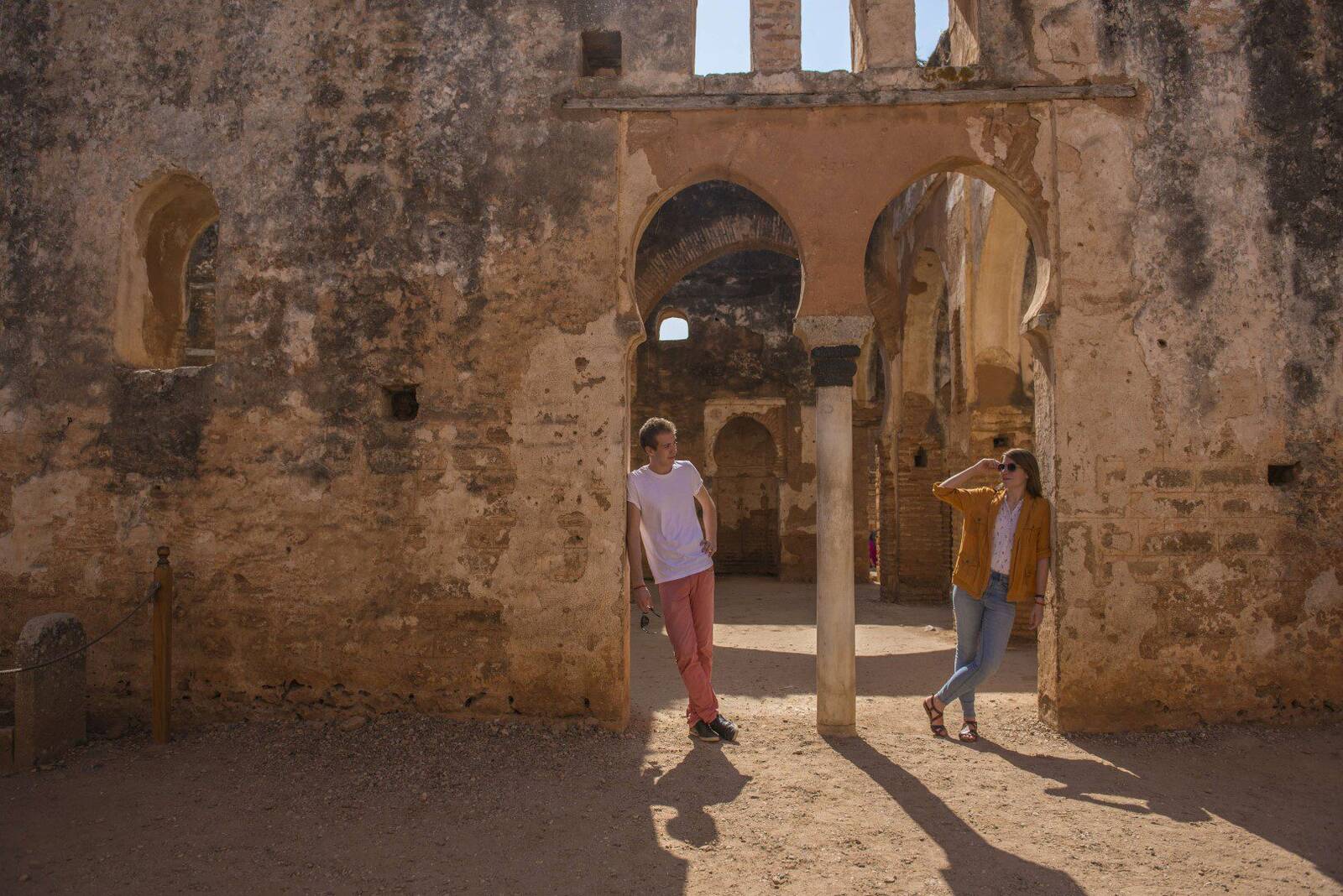 Couple standing apart beneath the ancient stone arches of the Chellah ruins, sunlight casting long shadows