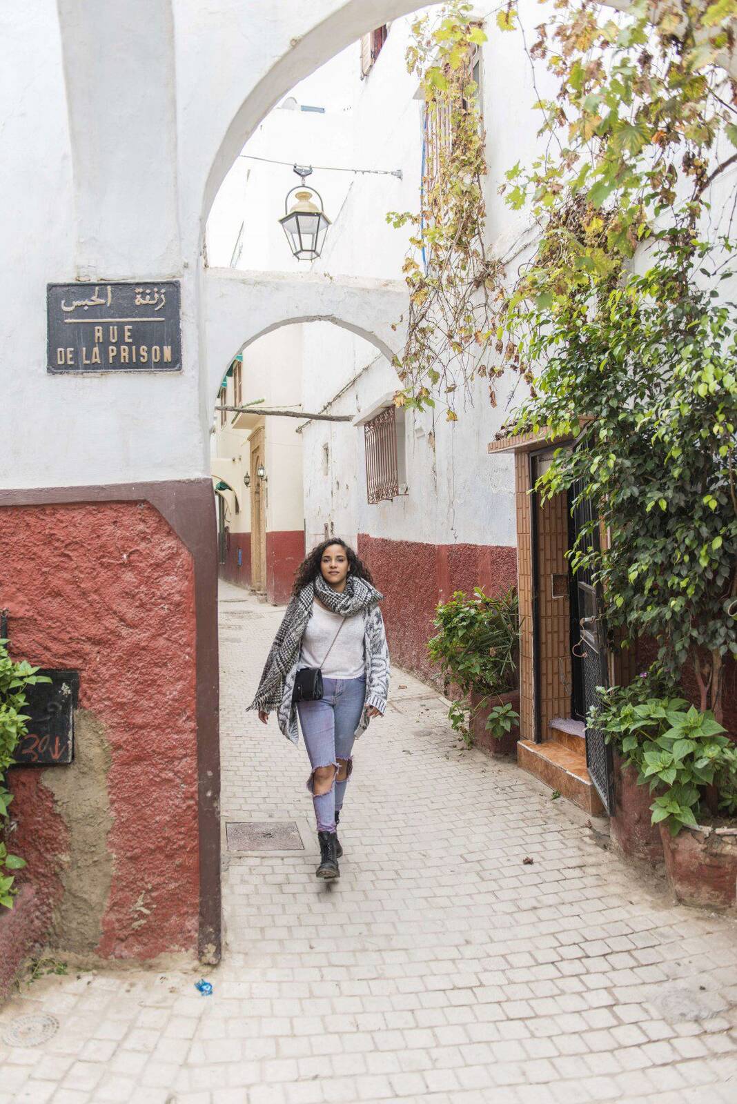 Woman in a scarf walking down a narrow cobblestone alley between red and white buildings with hanging plants in the Kasbah of the Oudayas