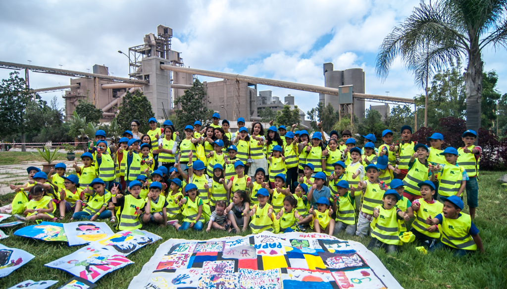 Group of workers in yellow safety helmets and vests posing outdoors at a cement plant, industrial towers in the background