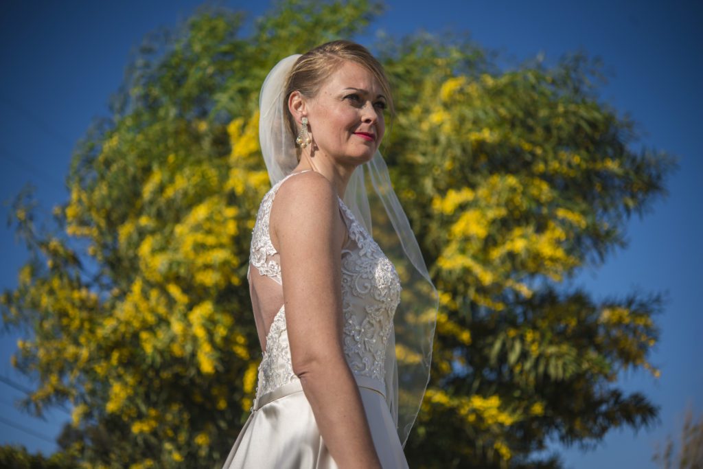 Bride in a white wedding gown and veil posing outdoors against a backdrop of yellow blossoming trees