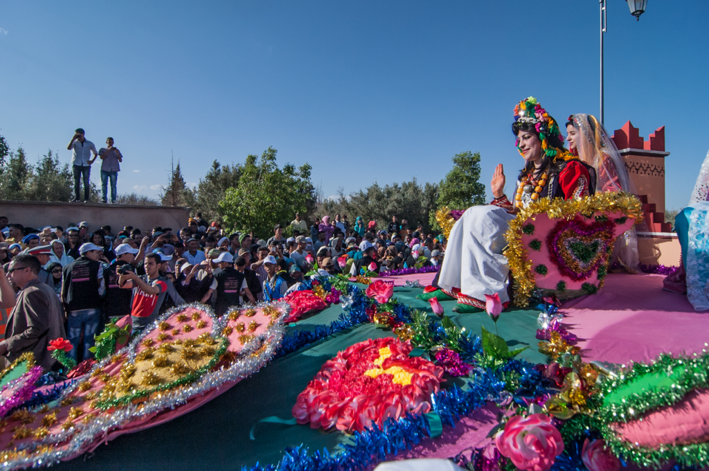 Colorful flower-covered parade float carrying performers in traditional dress, crowd watching at the Festival of Roses celebration