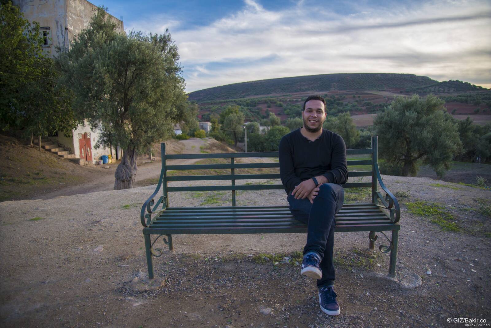 Man in a black sweater sitting on a green bench overlooking rolling hills and olive trees, GIZ youth employment program