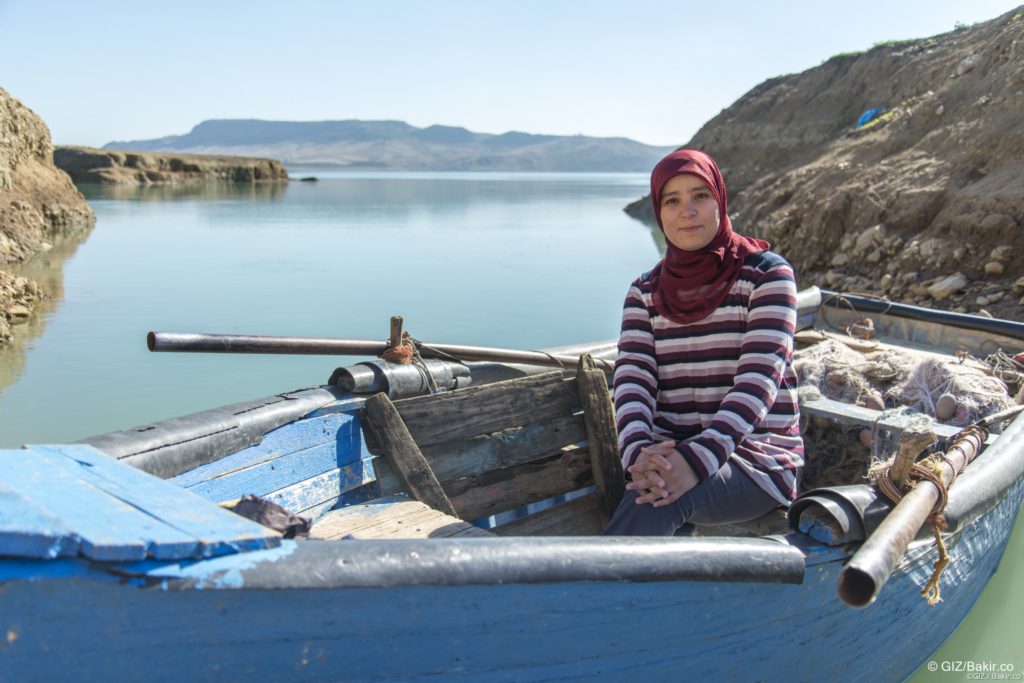 Woman in a maroon hijab and striped sweater sitting in a blue wooden rowboat on a calm lake, hands folded in her lap, hills in the background