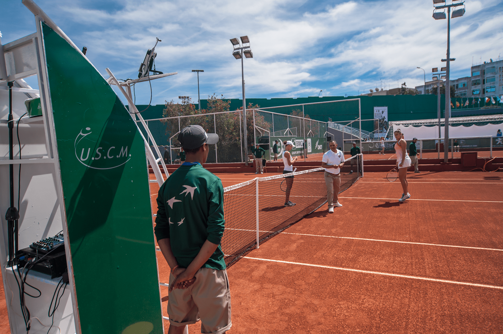 Man in a green U.S.C.M. polo standing courtside near a scoreboard while players gather at the net on a clay court