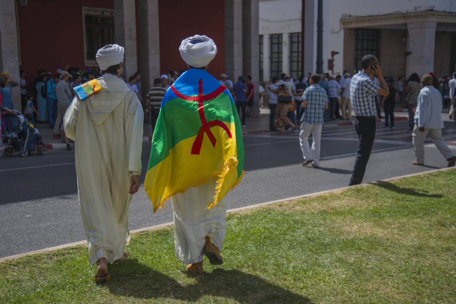 Two men seen from behind wrapped in an Amazigh flag walking across a grassy square toward a colonnaded building