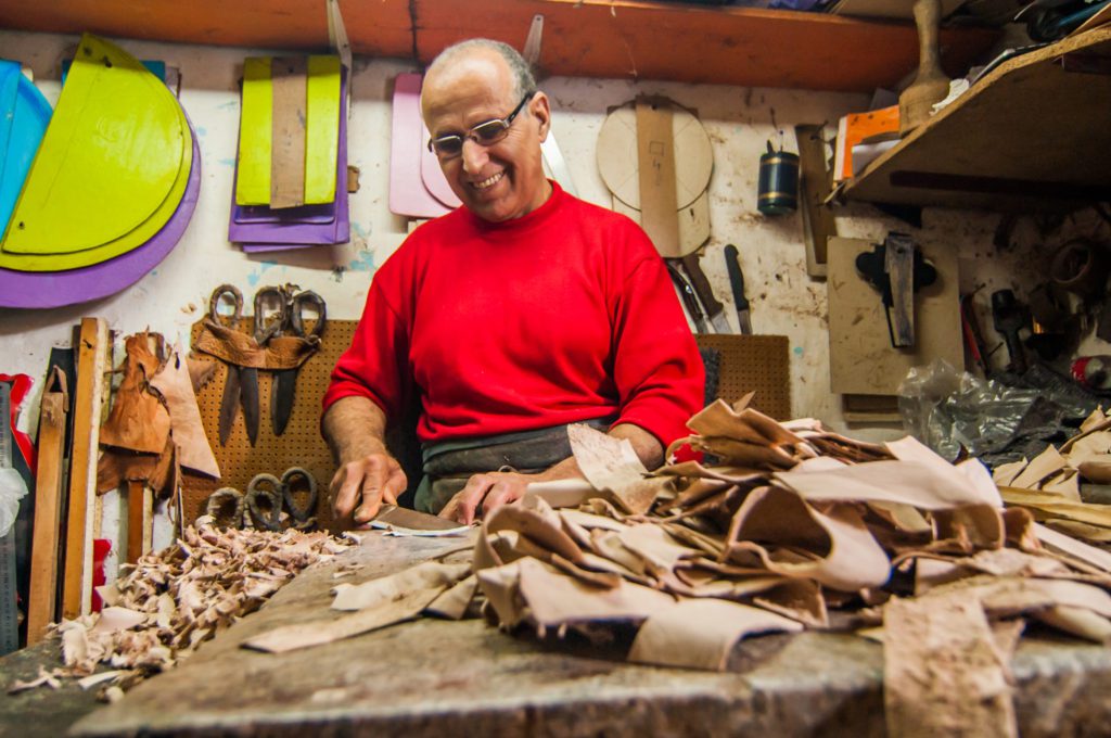 Craftsman in a red sweater working with leather hides and tools in a workshop, leather goods hanging on the wall