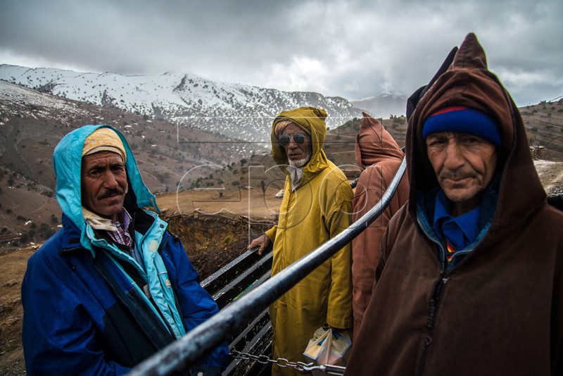 Two men wrapped in traditional wool hooded cloaks standing in a snowy mountain landscape