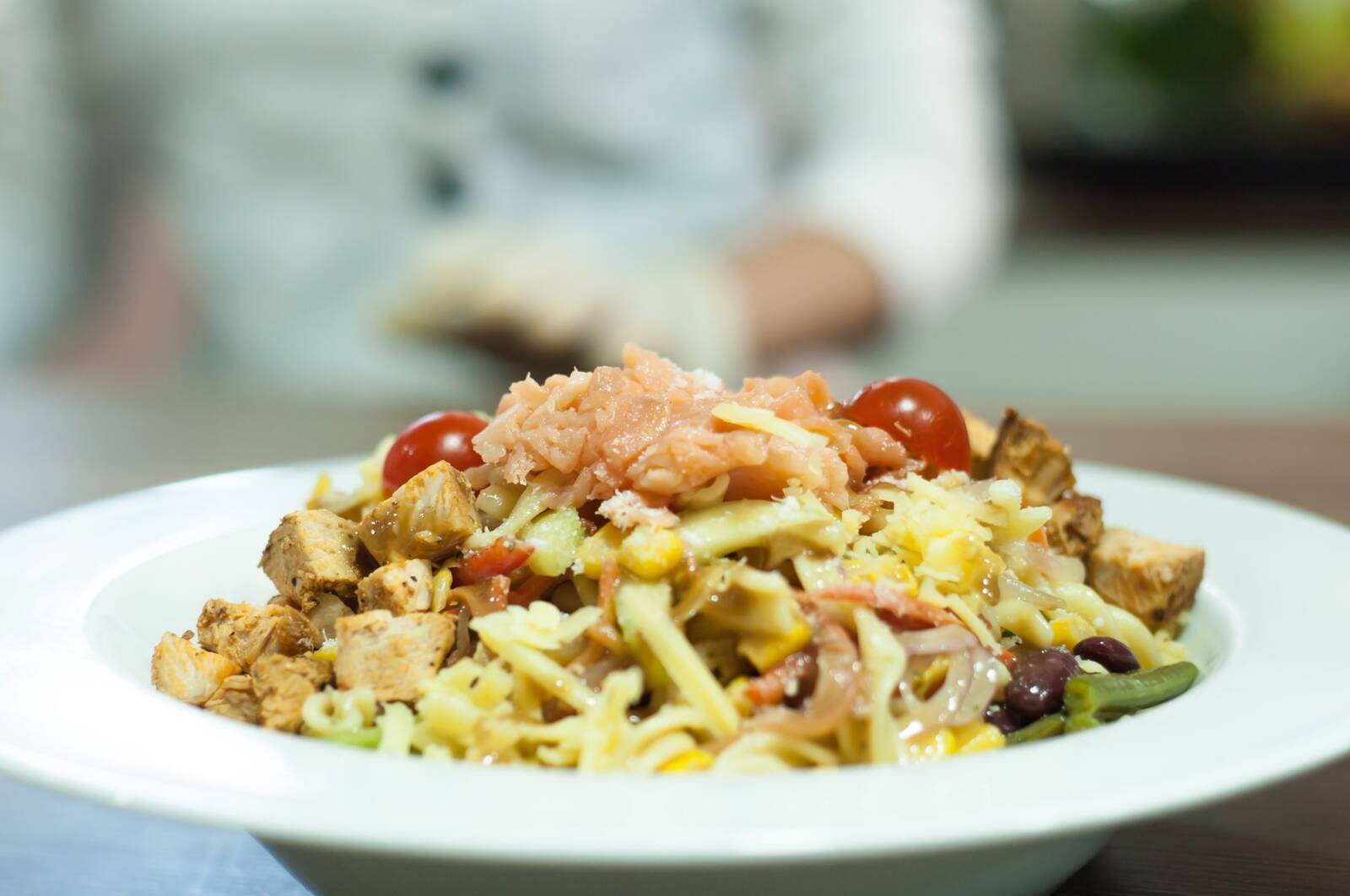Overhead shot of a chicken pasta salad with cherry tomatoes, corn, olives, and shredded meat in a white bowl, a chef in white blurred behind