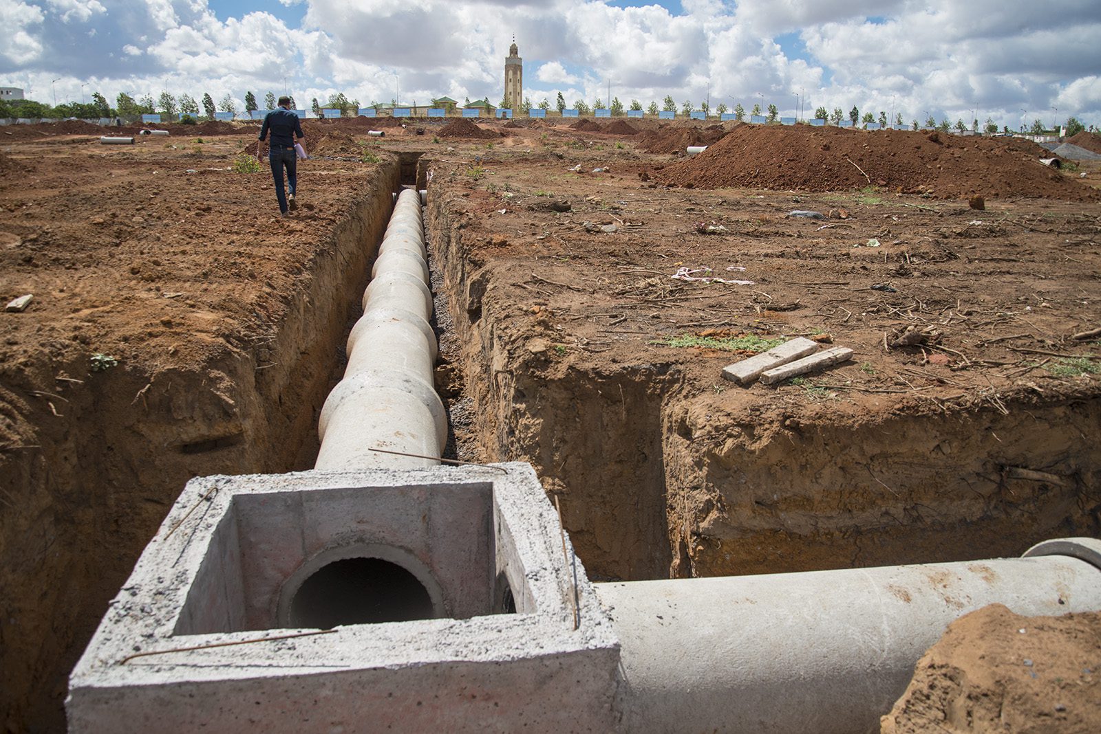 Concrete pipeline leading to a square manhole junction box in a trench, a worker walking along the edge toward a mosque minaret