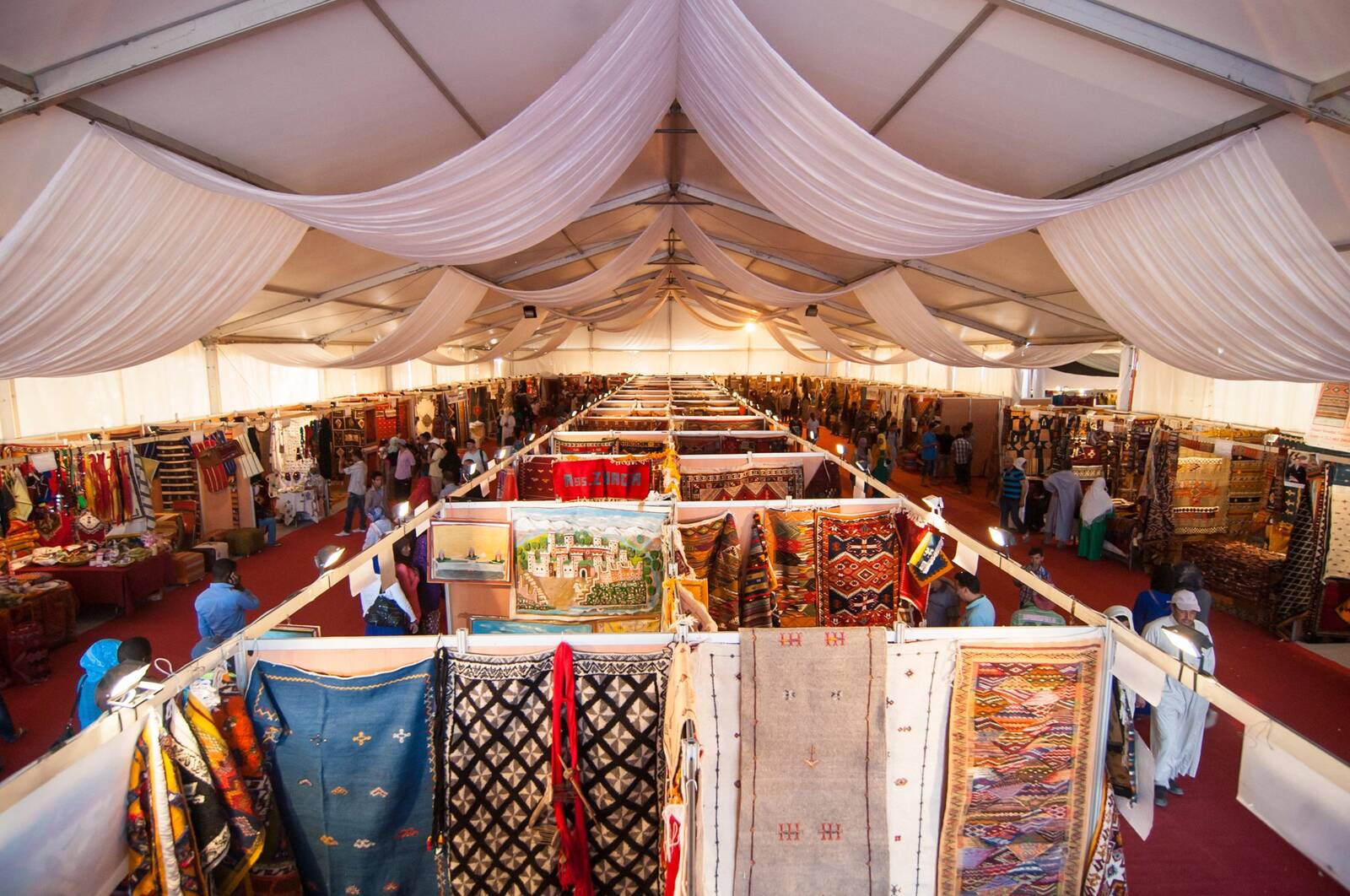 Wide interior view of a carpet festival hall under draped white fabric ceiling, rows of vendor stalls hung with rugs