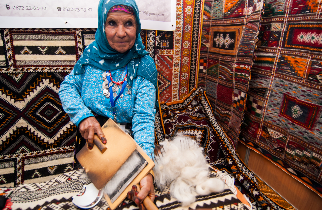 Woman in a blue headscarf demonstrating carpet weaving surrounded by colorful rugs at the Ouzguiti Carpet Festival