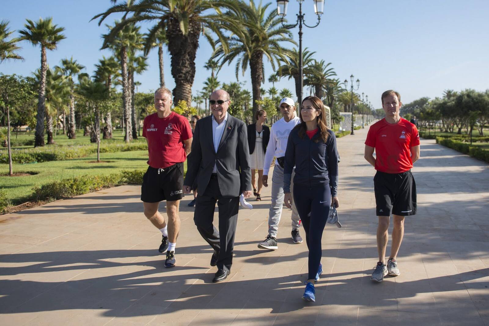 Group of people walking along a palm-tree-lined pathway, a man in a suit walking alongside others in athletic wear