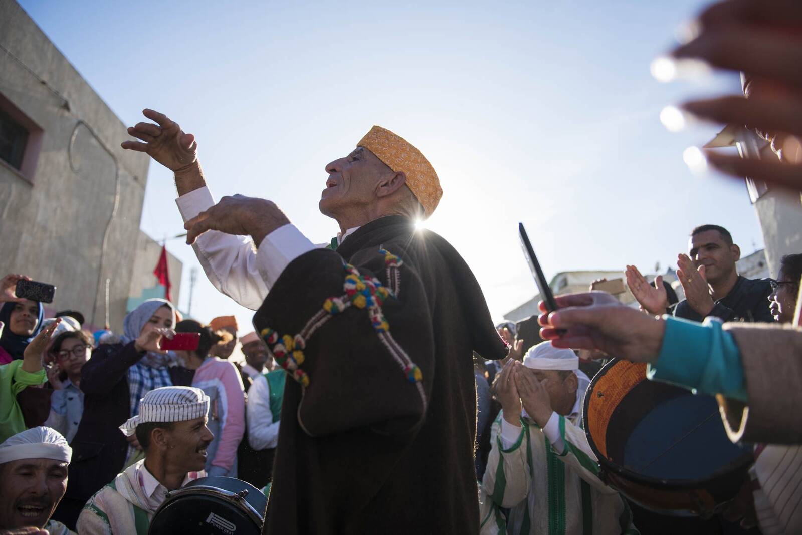 Crowd gathered outdoors at a religious moussem festival, a man in a white robe and turban raising his arm, backlit by sunlight