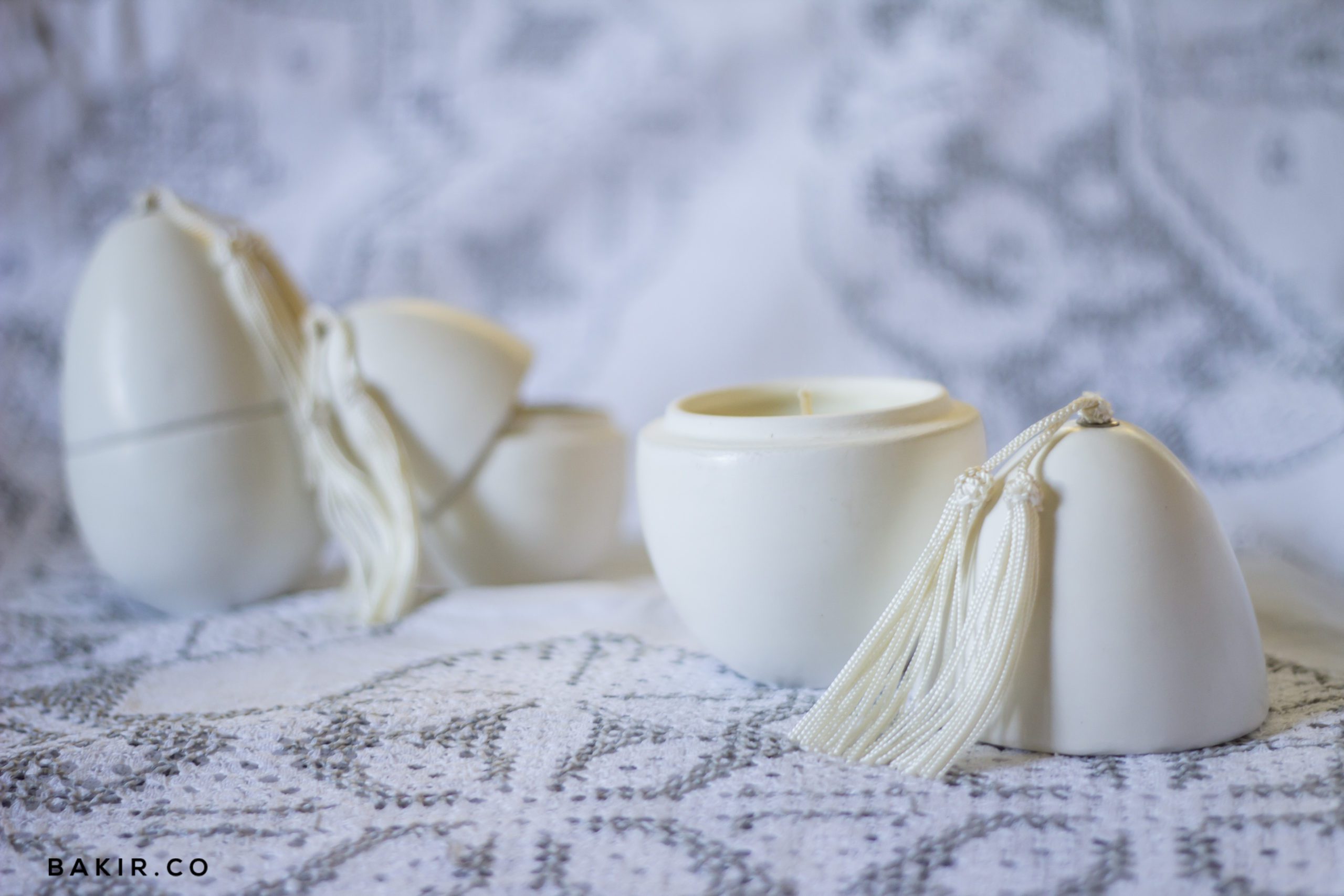 White ceramic candle jars with cream tassel ties arranged on an embroidered Moroccan-pattern fabric on an outdoor table