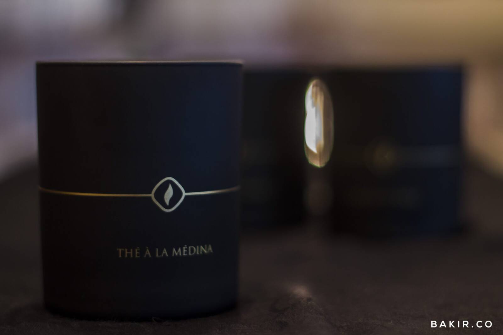 Black candle tin labeled "The a la Medina" with a gold flame logo, another tin blurred in the background