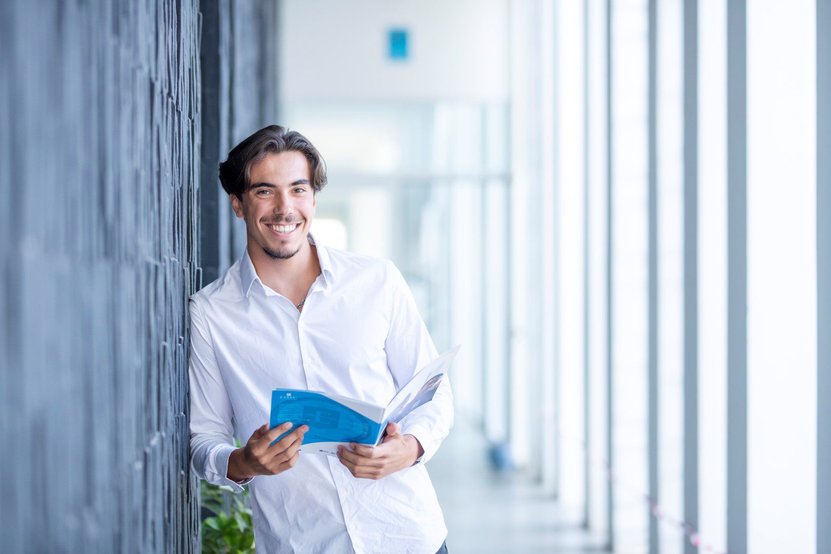 Young man leaning against a wall smiling while holding a brochure in a corridor