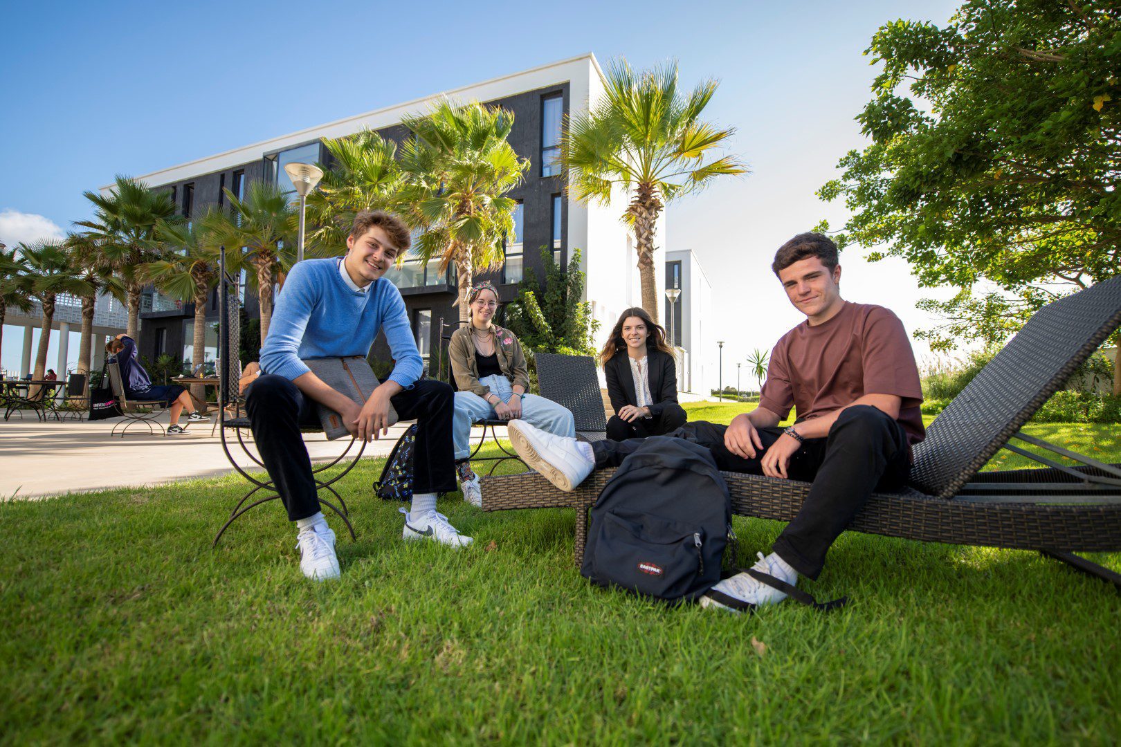 Four students relaxing on a lounge chair and bench on the lawn in front of the campus building