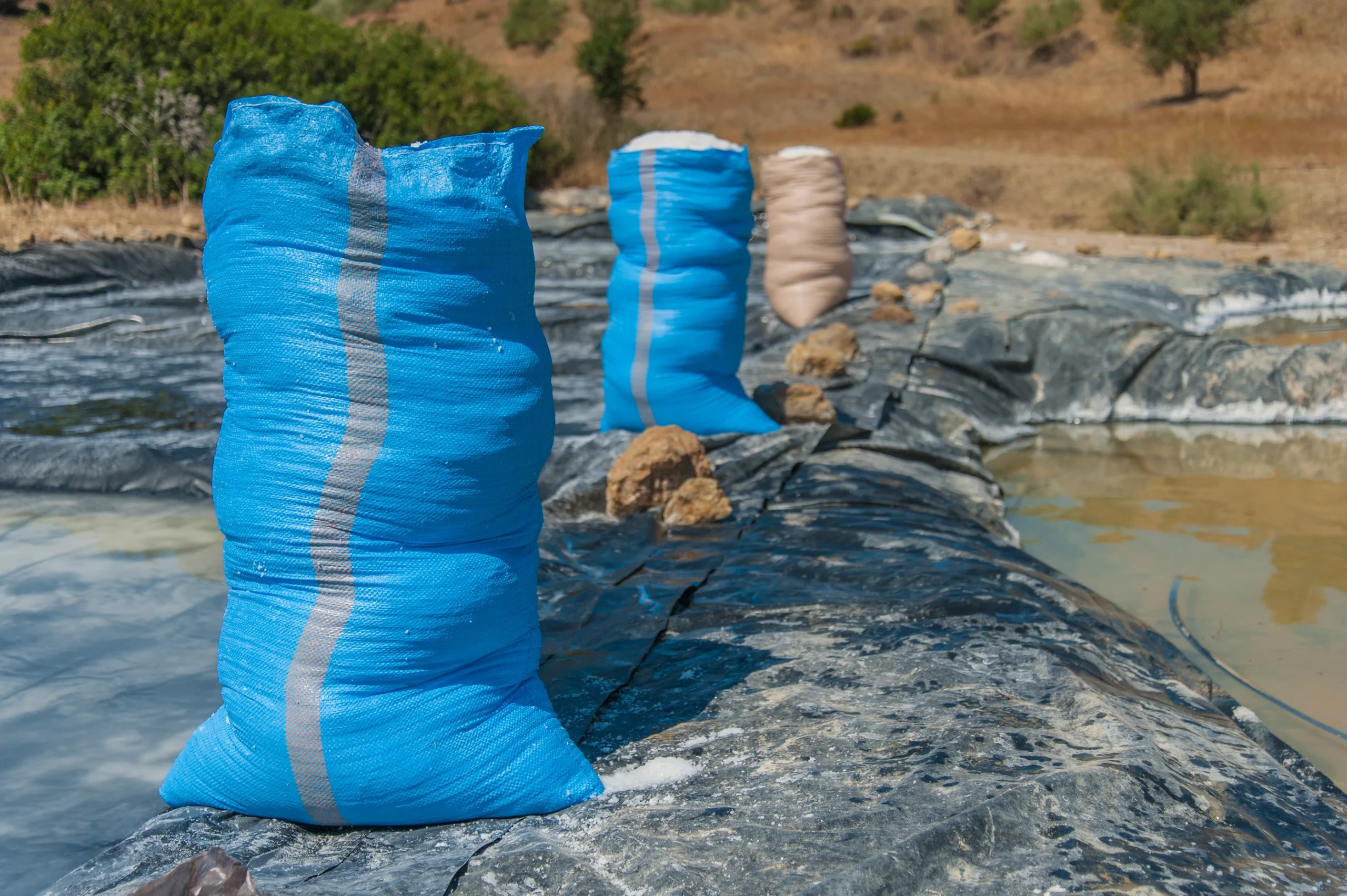 Blue and beige plastic sacks filled with salt standing along the edge of a stone salt-evaporation pond