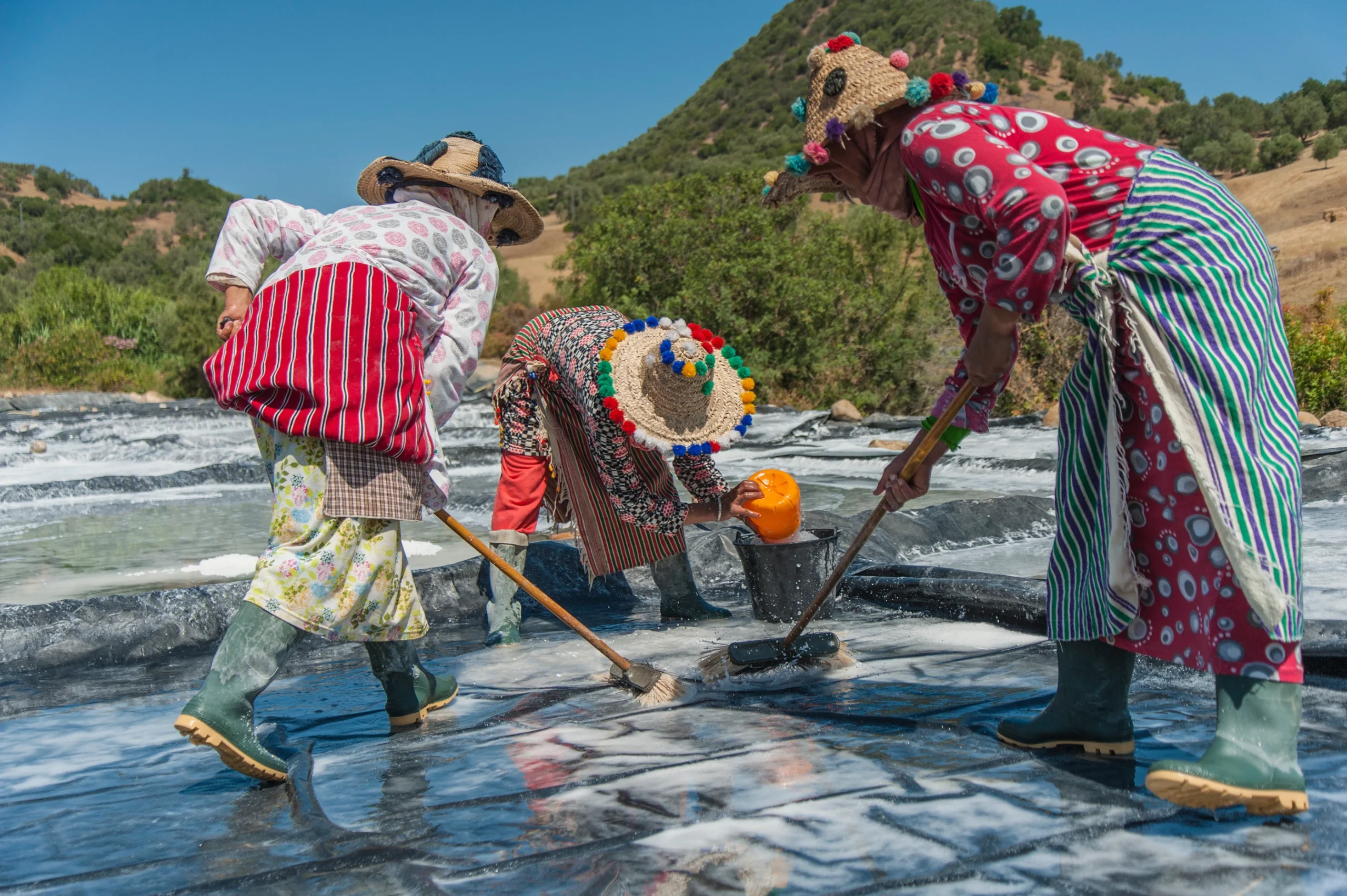 Three women in traditional dress and tasseled straw hats raking and hauling salt across black-lined evaporation ponds