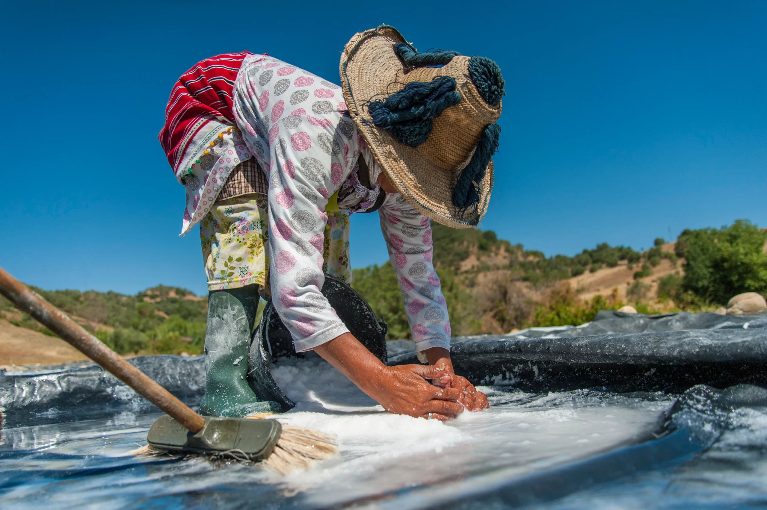 Woman in a floral robe and straw hat crouching to gather wet salt by hand at the edge of an evaporation pond